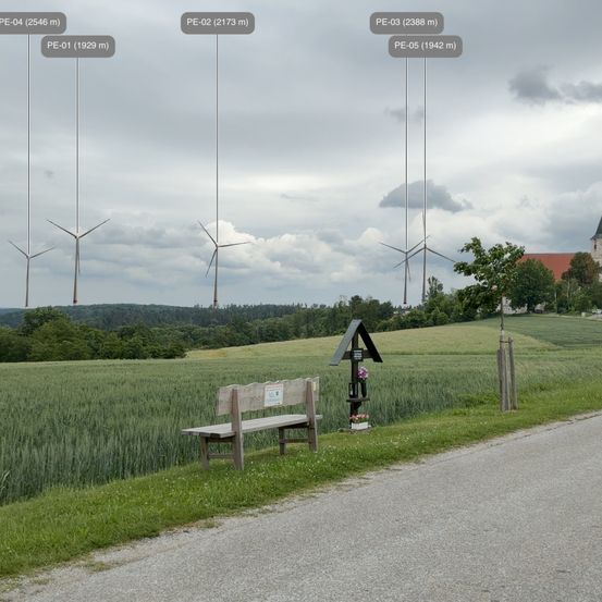 Eine ländliche Straße mit einer Holzbank und einer kleinen Struktur mit Blumen. In der Ferne befinden sich fünf Windturbinen mit Höhenmessungen. Der Himmel ist bewölkt und es gibt Bäume und ein Gebäude im Hintergrund.