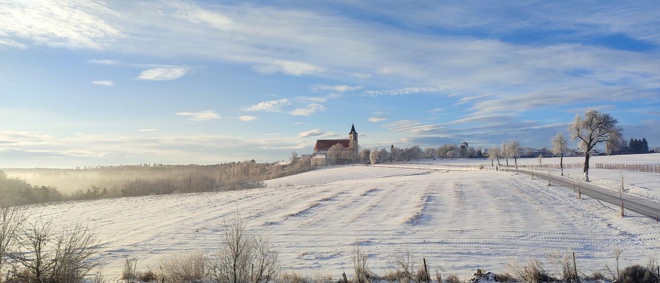 Eine Winterlandschaft mit einer Kirche, einem Turm und einem verschneiten Feld. Der Himmel ist blau mit verstreuten Wolken.