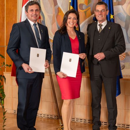 Drei Personen stehen zusammen, halten Urkunden in der Hand, in einer formellen Umgebung mit Fahnen und einer Wandmalerei im Hintergrund.