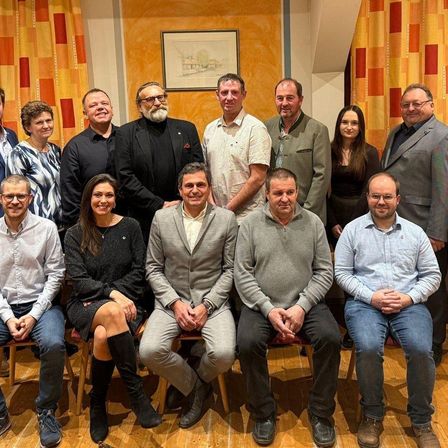 Eine Gruppe Erwachsener, darunter Männer und Frauen, posiert für ein Foto auf einem Holzboden. Sie sind feierlich gekleidet. Hinter ihnen befindet sich eine gelbe Wand mit Vorhängen und einem Bilderrahmen.