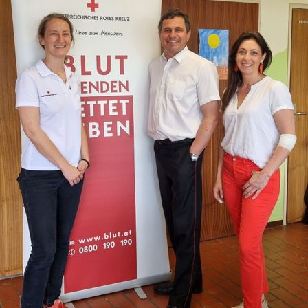 Drei Personen stehen vor einem rot-weißen Banner mit der Aufschrift 'BLUTRETTEN BEN'. Die Frau auf der linken Seite trägt ein weißes Shirt und blaue Jeans. Der Mann in der Mitte trägt ein weißes Hemd, schwarze Hose und eine Uhr. Die Frau auf der rechten Seite trägt ein weißes Hemd und rote Hose.
