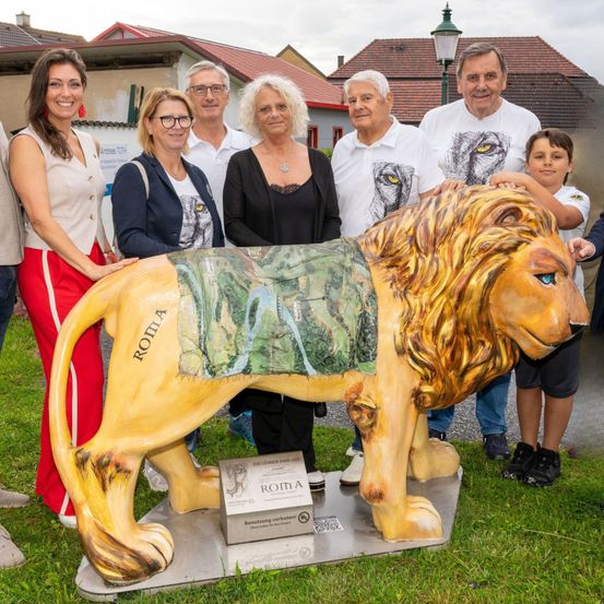 Eine Gruppe von Menschen posiert mit einer großen Löwenstatue mit dem Wort Roma auf der Seite, im Freien.