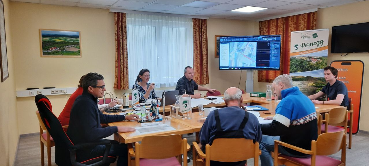 Eine Gruppe von Menschen ist in einem Besprechungsraum versammelt, wobei ein Mann auf einen großen Monitor zeigt, der eine Karte anzeigt. Der Raum hat einen Holztisch, Stühle und verschiedene Büromaterialien. Der Mann trägt ein schwarzes Hemd, während die Frau eine blaue und weiße Bluse hat.