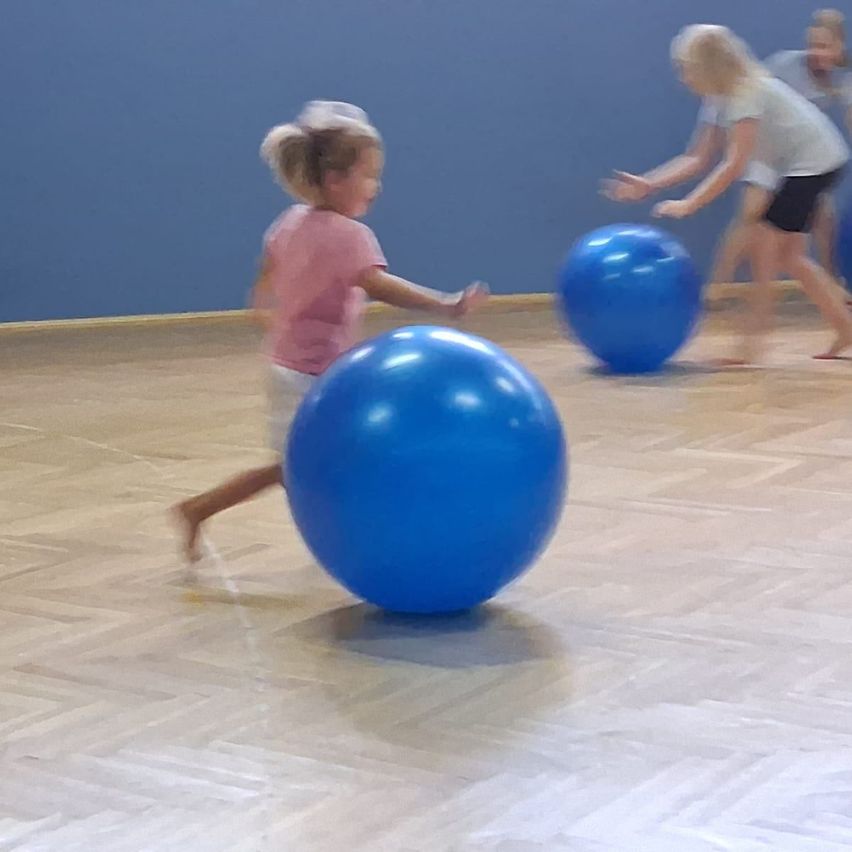 Einige Kinder spielen mit großen blauen Übungsbällen in einem Fitnessstudio. Ein Kind läuft auf einen Ball zu.