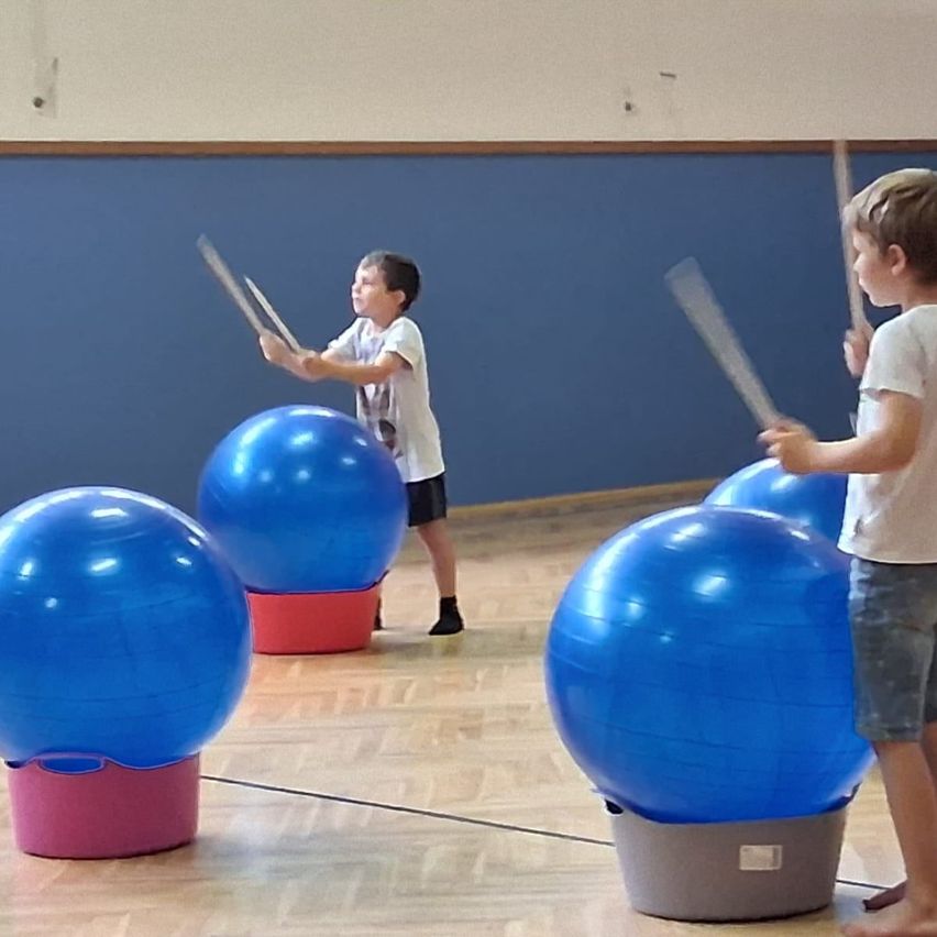Zwei Kinder spielen mit großen blauen Übungsbällen auf einem Holzboden. Sie halten Stöcke und stehen neben den Bällen.