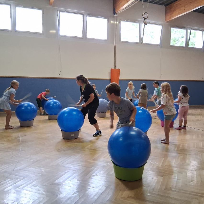 In einer Turnhalle spielen Kinder mit großen blauen Bällen, einige stehen auf dem Boden und andere auf grauen Sockeln. Eine Frau steht neben einem blauen Ball.