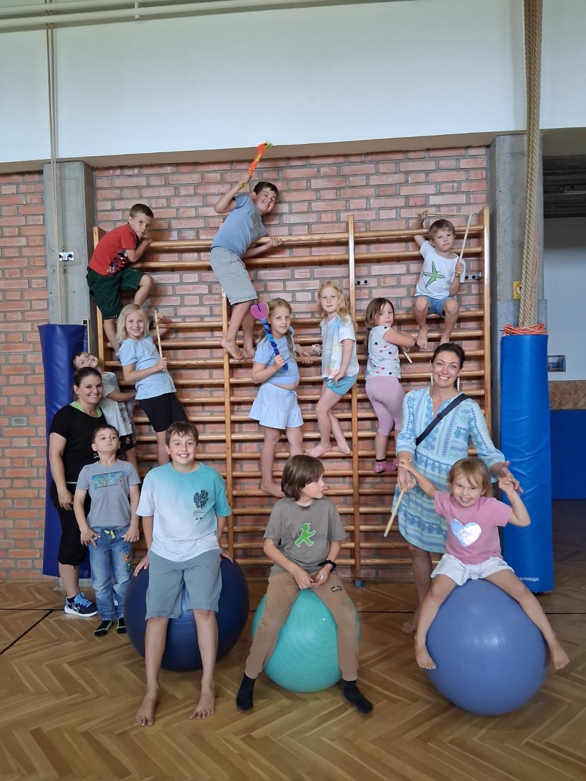 Eine Gruppe von Kindern und Erwachsenen befindet sich in einer Turnhalle. Einige Kinder sind auf einer Kletterleiter, während andere auf Gymnastikbällen sitzen. Die Erwachsenen lächeln und stehen hinter den Kindern.