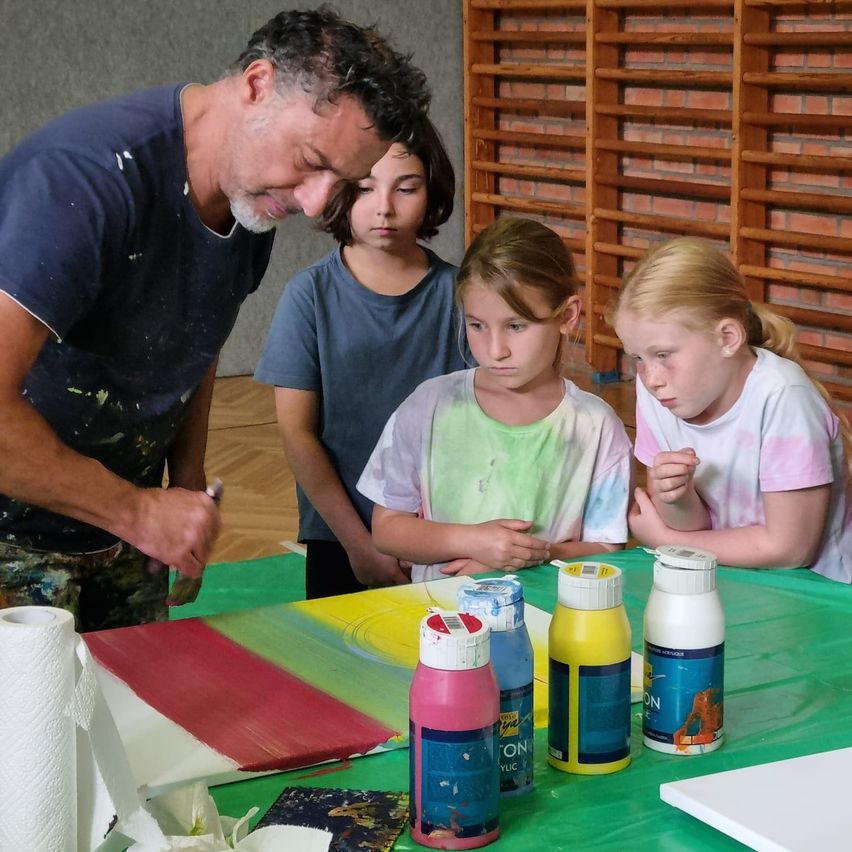 Ein Mann malt ein großes Leinwandbild, während drei junge Mädchen beobachten. Sie stehen um einen Tisch, der mit Farbflaschen und einem Papierhandtuch bedeckt ist.