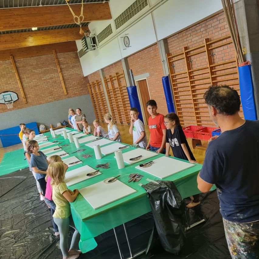 In einer Turnhalle stehen Kinder und ein Mann an einem langen Tisch, der mit grünem Tuch bedeckt ist, mit Malutensilien. Der Raum hat Ziegelwände, Holzbalken und Geräte.