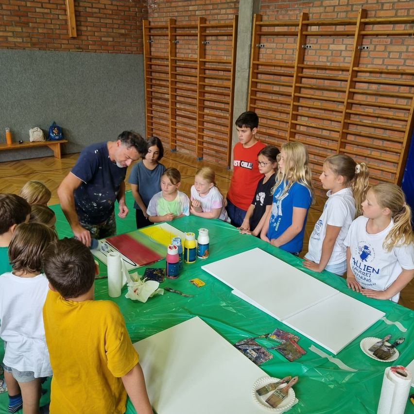 Eine Gruppe von Kindern und einem Erwachsenen versammelt sich um einen mit einem grünen Tuch bedeckten Tisch. Der Tisch hat Malutensilien und Papiere. Der Erwachsene steht und scheint die Kinder zu instruieren. Die Kinder schauen den Erwachsenen an und einige halten Malpinsel.