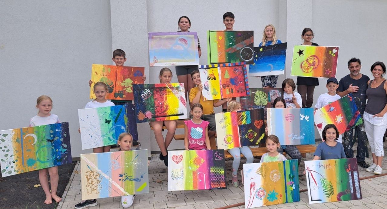 Eine Gruppe von Kindern steht und sitzt auf einer Bank und hält bunte Gemälde hoch. Einige Gemälde haben Text wie 'enjoy' und 'family'. Die Kinder lächeln und posieren für ein Foto.