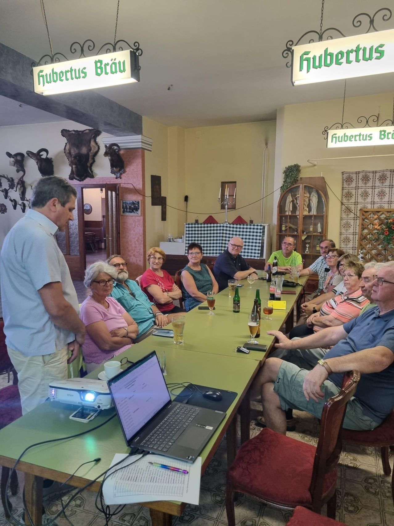 Eine Gruppe von Menschen sitzt um einen Tisch in einem Raum. Ein Mann steht und spricht. Ein Laptop, ein Projektor und Tassen stehen auf dem Tisch. Flaschen und Gläser sind auf dem Tisch.