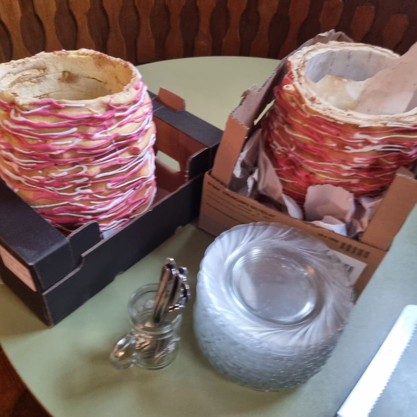 Zwei Kartons mit rosa und weißen spiralförmigen Objekten stehen auf einem Tisch. In der Nähe befindet sich eine Glasvase, ein kleines Glas mit Besteck und ein Messer.