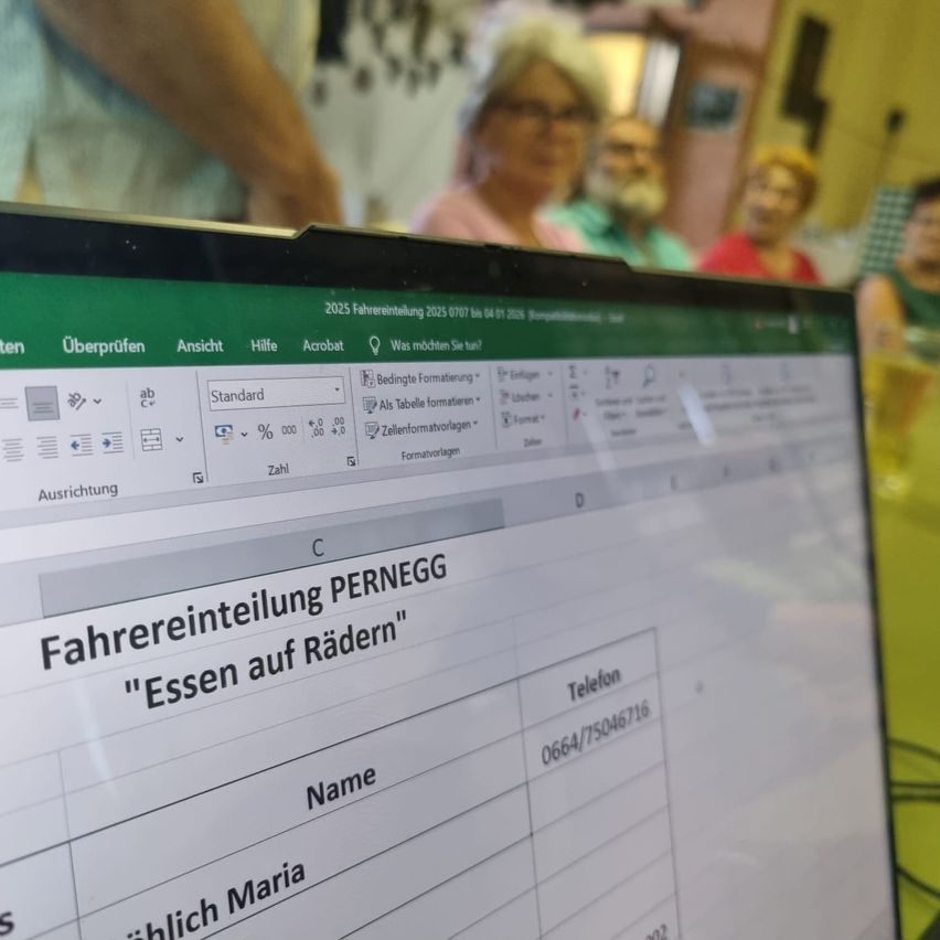 Ein Computerbildschirm zeigt ein Tabellenkalkulationsprogramm mit der Überschrift 'Fahrereinteiung PERNEGG'. Darunter steht 'Essen auf Rädern' mit dem Kontaktnamen Maria und der Telefonnummer 0664/75046716. Leute sitzen hinter dem Computer.