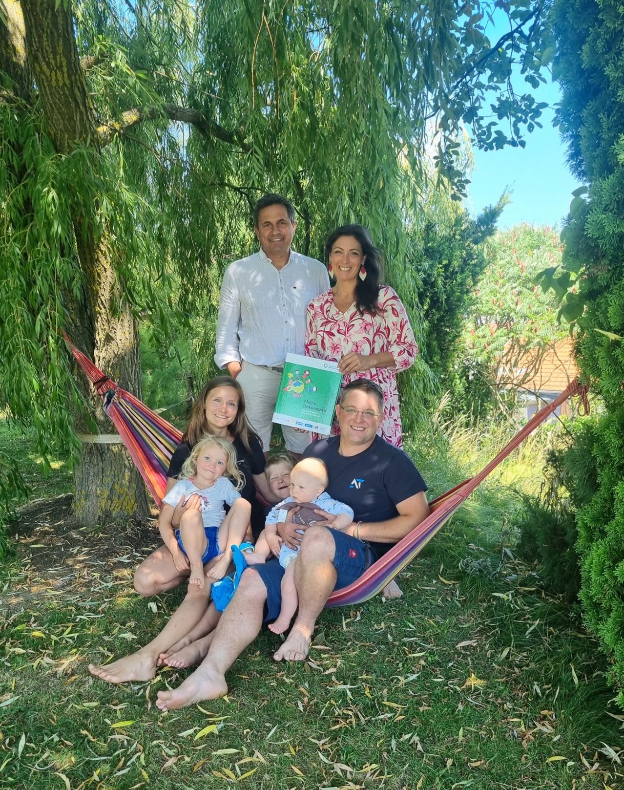 Eine Familie mit sechs Personen sitzt in einer Hängematte und hält ein grünes Zertifikat. Ein Erwachsener steht lächelnd hinter ihnen. Die Umgebung ist ein Garten mit Bäumen und Gras.