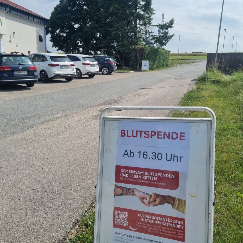 Ein Schild auf Deutsch am Straßenrand mit 'Blutspende' und einem QR-Code. Autos sind vor einem Gebäude geparkt.