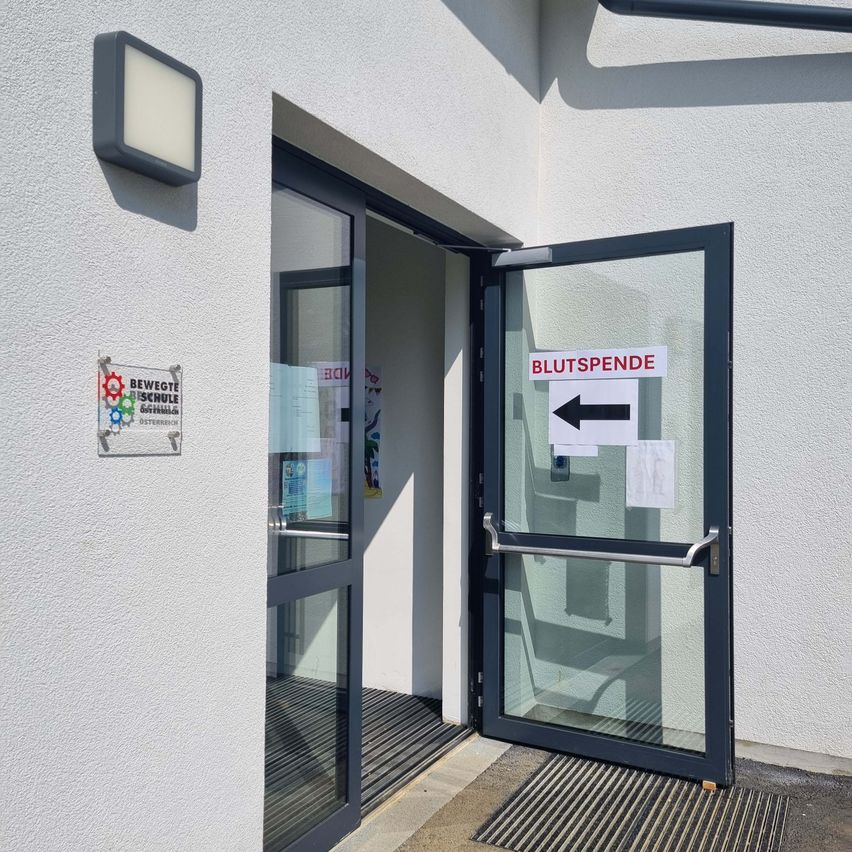 Eine offene Tür führt zu einem Gebäude mit einem Schild, das auf Blutspende hinweist. Es gibt ein weiteres Schild an der Wand und eine Lampe, die darüber montiert ist.