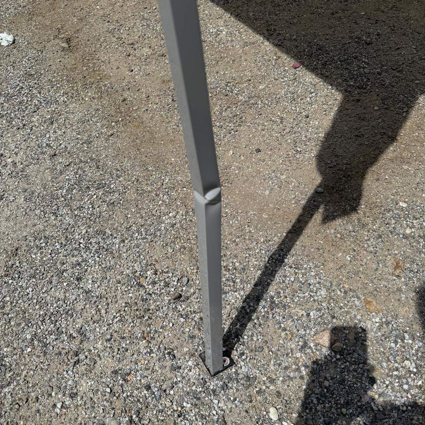 Eine Nahaufnahme eines grauen Metallpfostens auf einer Kiesoberfläche mit einer gebogenen Stelle am unteren Ende. Der Schatten einer Person fällt auf den Boden.
