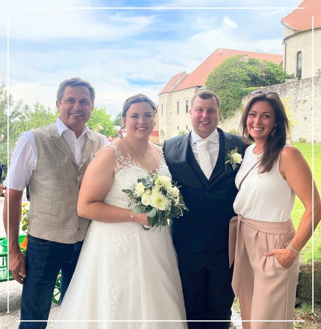 Ein frisch verheiratetes Paar steht mit zwei anderen lächelnd vor einem Schloss mit rotem Dach. Der Bräutigam trägt einen schwarzen Anzug und die Braut ein weißes Kleid.