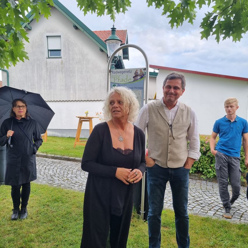 Drei Menschen stehen vor einem Gebäude, eine Frau hält einen Regenschirm. Hinter ihr stehen eine weitere Frau und ein Mann. Sie lächeln und posieren für ein Foto. Ein Schild ist an dem Pfosten befestigt.
