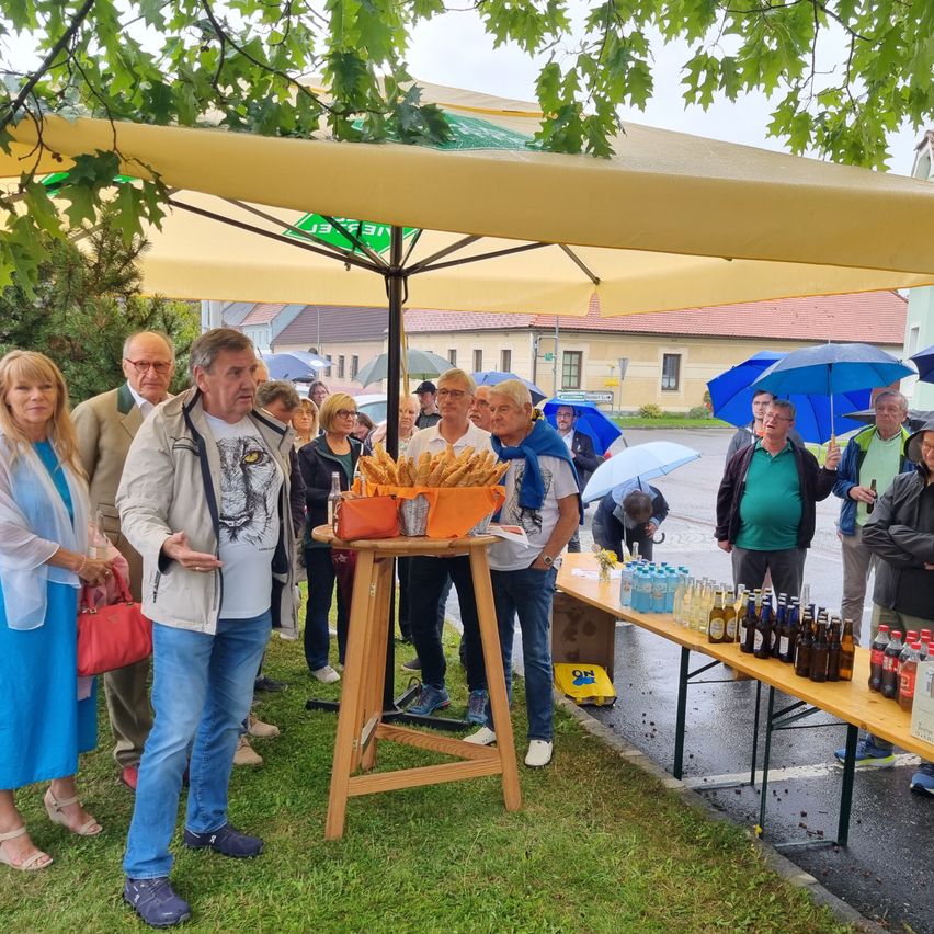 Eine Gruppe von Menschen versammelt sich unter einem großen Regenschirm. Einige halten Regenschirme. Ein Mann spricht, während ein anderer neben einem Tisch mit Essen steht. Flaschen und Dosen sind auf einem Tisch in der Nähe.