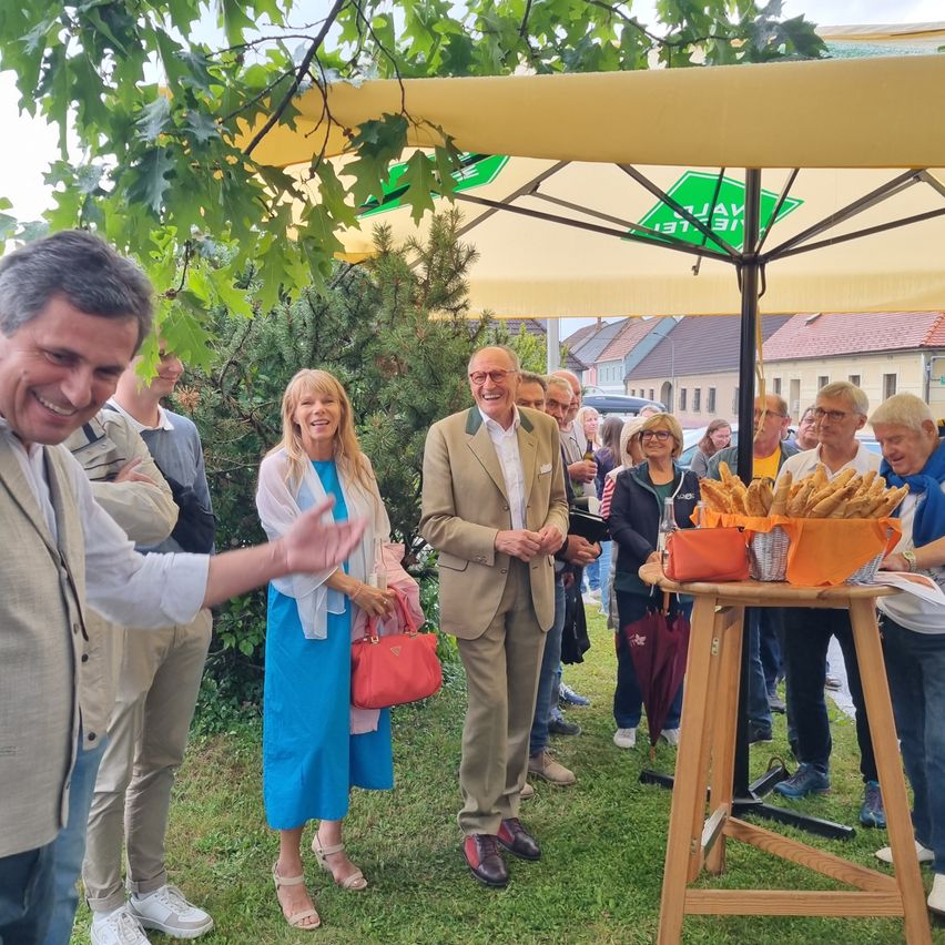 Mehrere Menschen versammeln sich unter einem gelben Regenschirm im Freien. Ein Mann lächelt, während ein anderer Mann gestikuliert. In der Nähe steht ein Tisch mit Körben mit Brot.