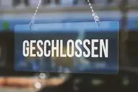 Ein blaues Schild mit weißem Text, das 'GESCHLOSSEN' liest, hängt an einer Kette. Es befindet sich vor einem unscharfen Hintergrund, möglicherweise einem Schaufenster.