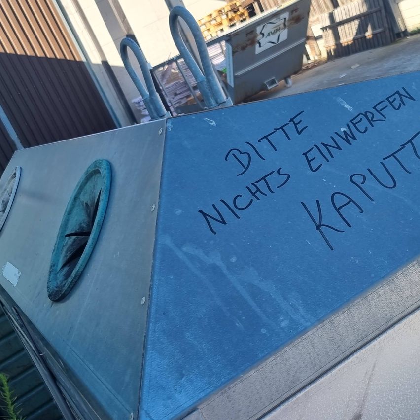 Ein blauer Container mit Graffiti, das 'BITTE EINWERFEN NICHTS KAPUTT' besagt, steht draußen in der Nähe eines Gebäudes und eines Metallzauns.