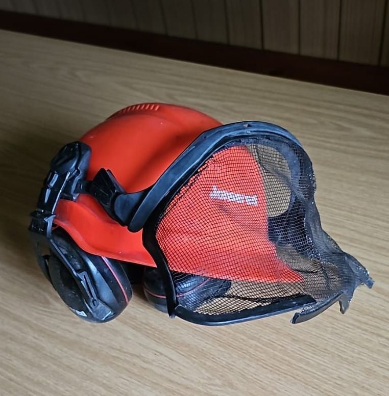 Ein roter Helm mit Netzgesichtsschutz liegt auf einer Holzoberfläche. Der Helm hat einen schwarzen Riemen.