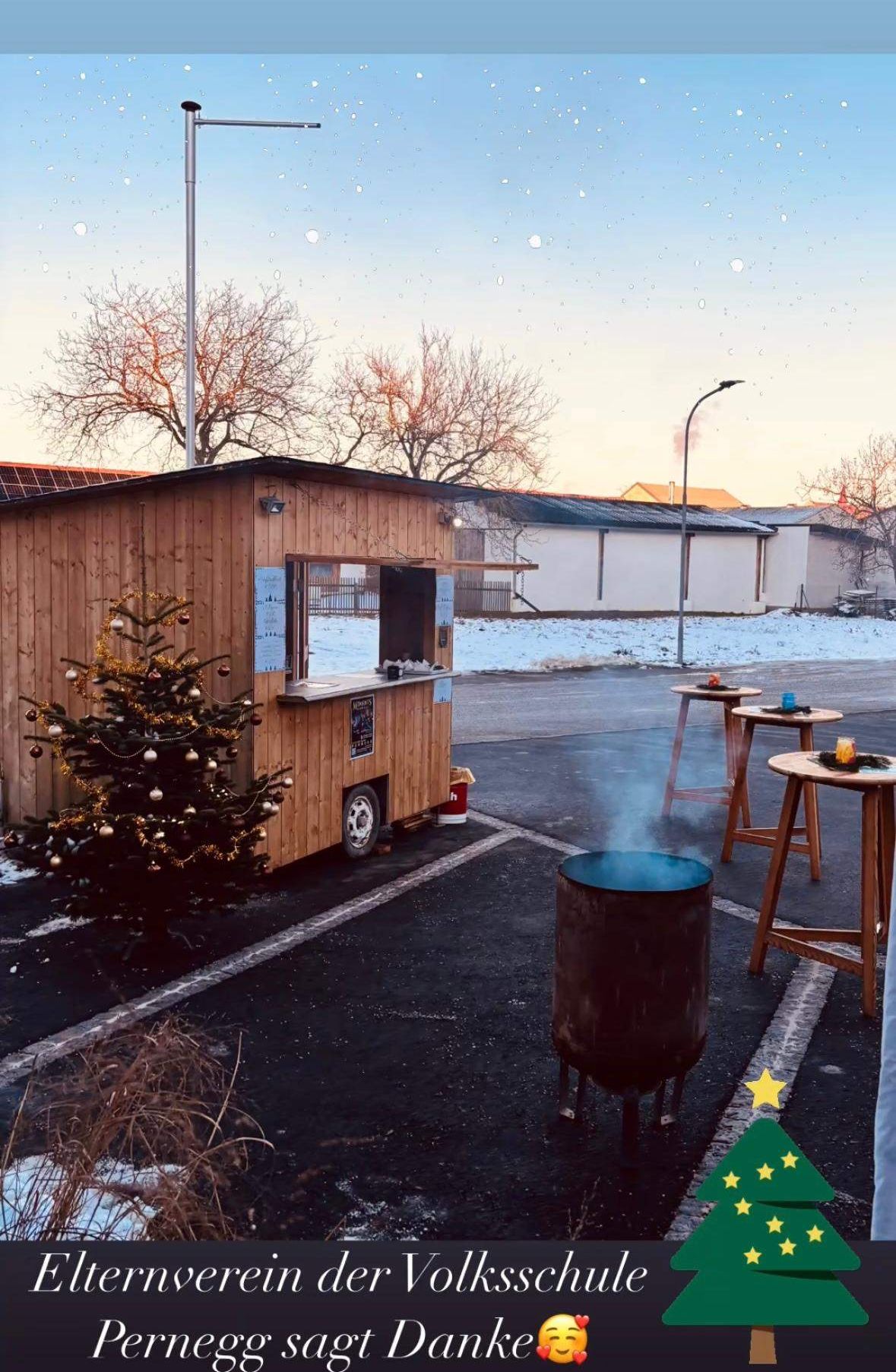 Ein Holzimbisswagen mit einem Weihnachtsbaum ist in einem verschneiten Bereich geparkt, mit einem Fass, das Rauch abgibt, zwei Tischen und einem Pfosten.