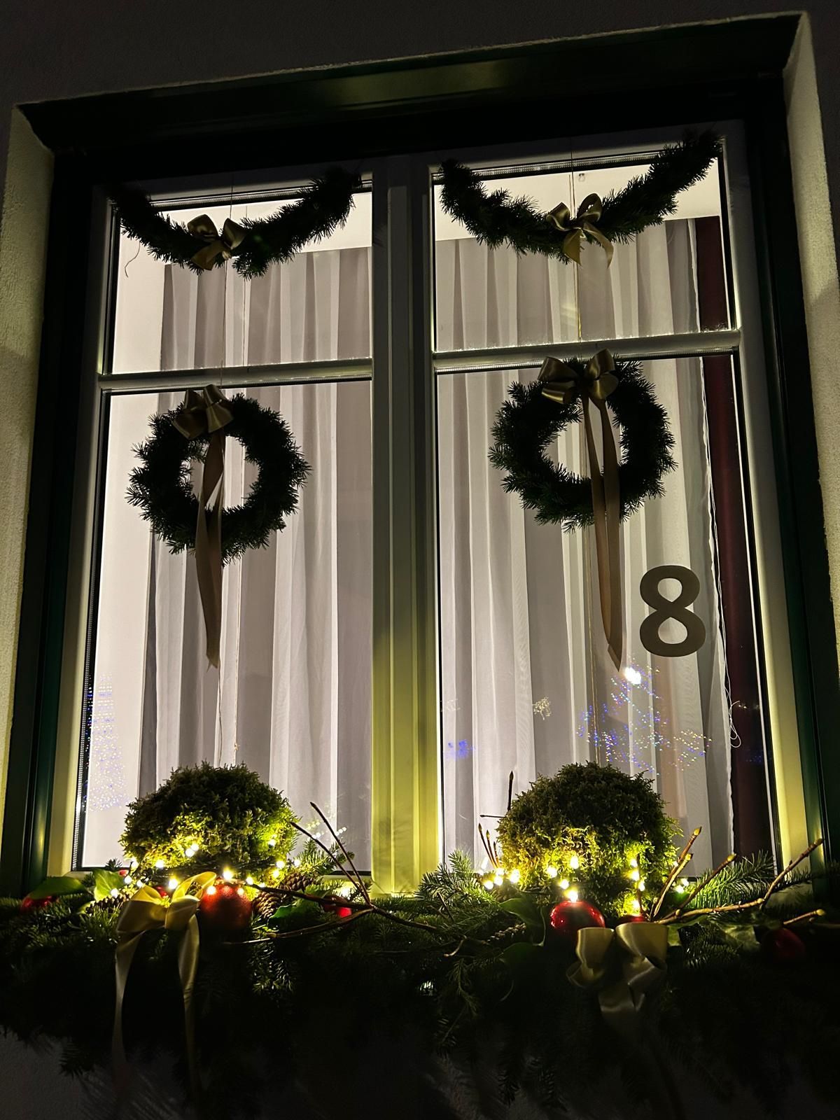 Two windows decorated for Christmas with wreaths, garlands, and a number 8. The windows have white curtains and are lit up with lights.