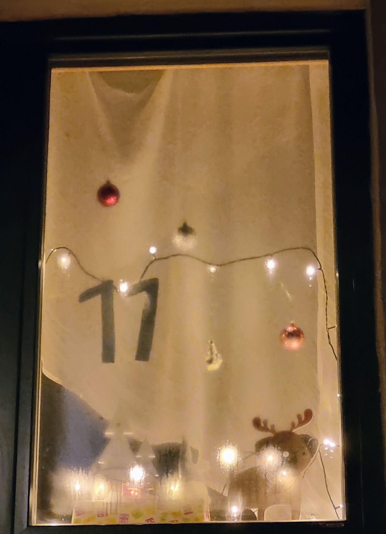 Ein Fenster zeigt eine Weihnachtsszene mit der Nummer 17 und festlichen Lichtern. Die Nummer ist groß, und mehrere Ornamente hängen von den Lichtern. Der Hintergrund ist unscharf.