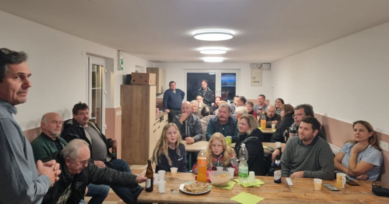 Eine Gruppe von Menschen sitzt an Tischen in einem Raum mit Flaschen und Essen. Zwei Frauen stehen sich mit Lächeln im Gesicht gegenüber.