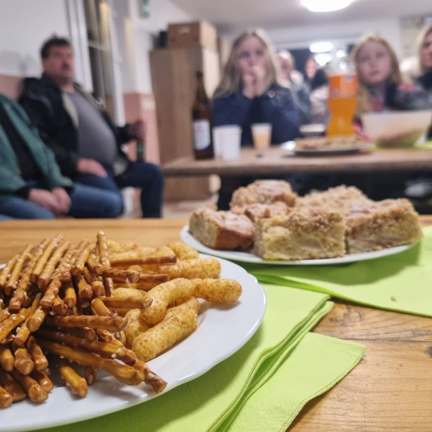 Eine Gruppe von Menschen sitzt an einem Tisch, mit Snacks auf einem Teller im Vordergrund, einschließlich Brezeln und Käsepuffs. Dahinter sitzen weitere Menschen und ein Tisch mit Essen und Getränken.