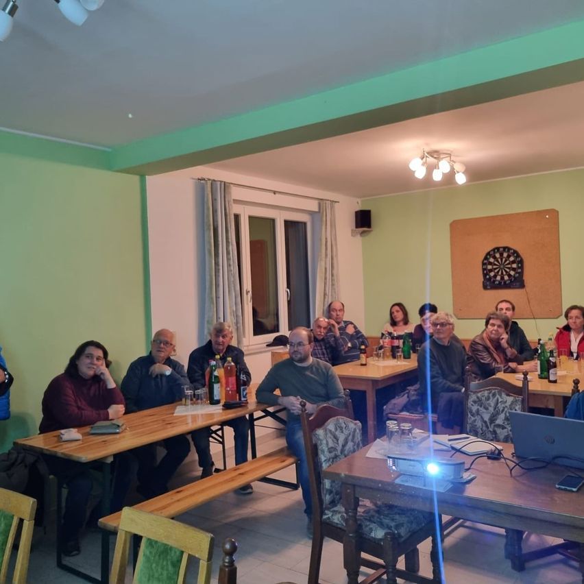 Eine Gruppe von Menschen, die um Tische herum in einem Raum mit grünen Wänden und einem Dartboard an der Wand sitzen. Flaschen und Gläser auf dem Tisch.