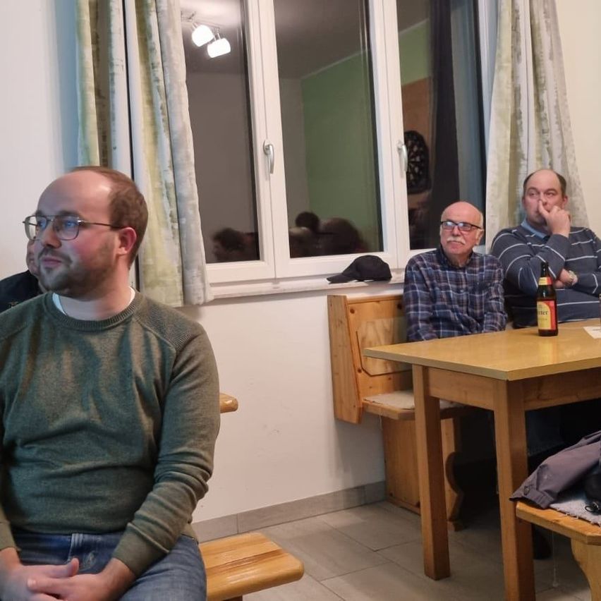 Drei Männer sitzen an einem Holztisch in einem Raum mit Fenster und Vorhängen. Einer trägt eine Brille, und ein anderer hat eine Bierflasche auf dem Tisch.