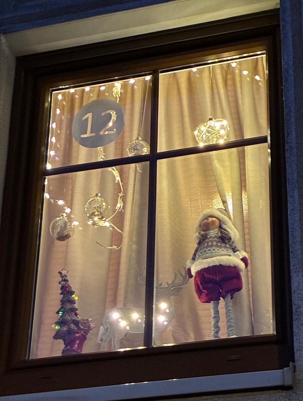 Ein Fensterdisplay mit einem Weihnachtsbaum, einer Puppe in Winterkleidung und Dekorationen, alles beleuchtet von Lichterketten, mit der Nummer 12 sichtbar am Fenster.