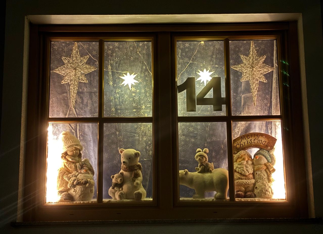 Ein Fensterdisplay mit Weihnachtsdekorationen, darunter eine Nummer 14 und Ornamente wie ein Stern, ein Bär, ein Schneemann und ein Rentier. Das Fenster ist beleuchtet und zeigt die Dekorationen.