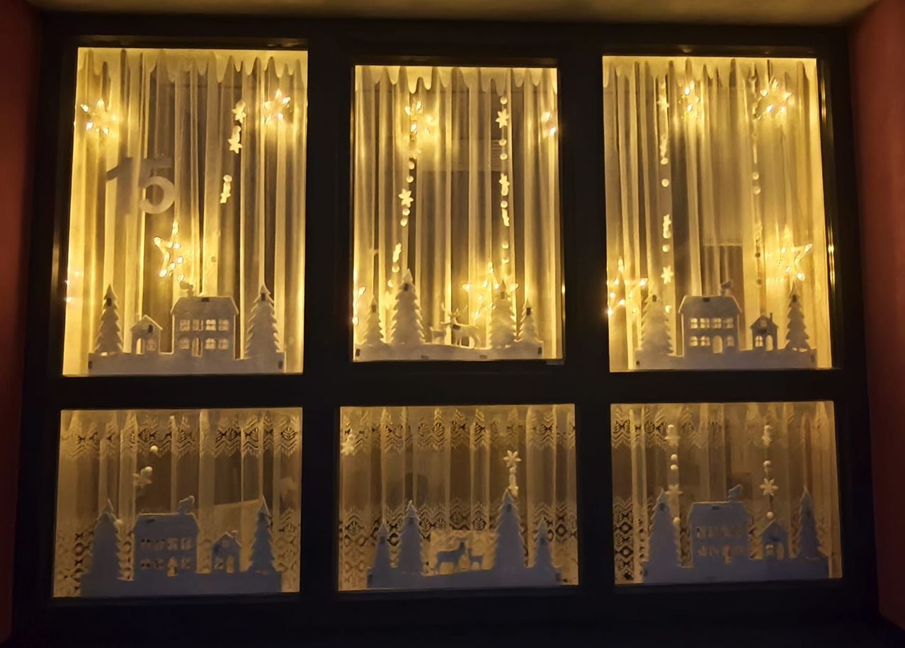 Eine Reihe von festlichen Fensterdekorationen mit weißen Silhouetten von Weihnachtsbäumen, Häusern und Sternen. Die Lichter erzeugen ein warmes, einladendes Leuchten.