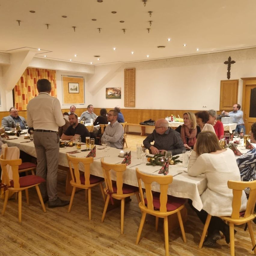 Eine Gruppe von Menschen sitzt an langen Tischen in einem Speisesaal. Ein Mann steht vor ihnen, möglicherweise hält er eine Rede. Die Tische sind mit Gläsern, Tellern und Weingläsern gedeckt. Der Raum hat Holzböden und Wände mit Bilderrahmen und einem Kreuz.