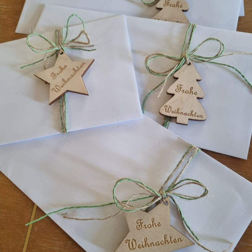 Mehrere weiße Umschläge mit grünem Faden und Holzanhängern liegen auf einer Holzoberfläche. Die Anhänger sind ein Stern und ein Baum, beide mit der Aufschrift 'Frohe Weihnachten.'