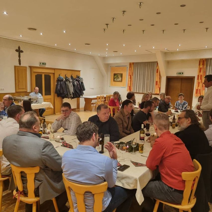 Eine Gruppe von Männern sitzt um einen langen Tisch in einem Esszimmer herum und führt ein Gespräch. Der Tisch ist mit Weingläsern, Flaschen und Servietten gedeckt. Der Raum hat Holzstühle und eine warme Beleuchtungseinrichtung.