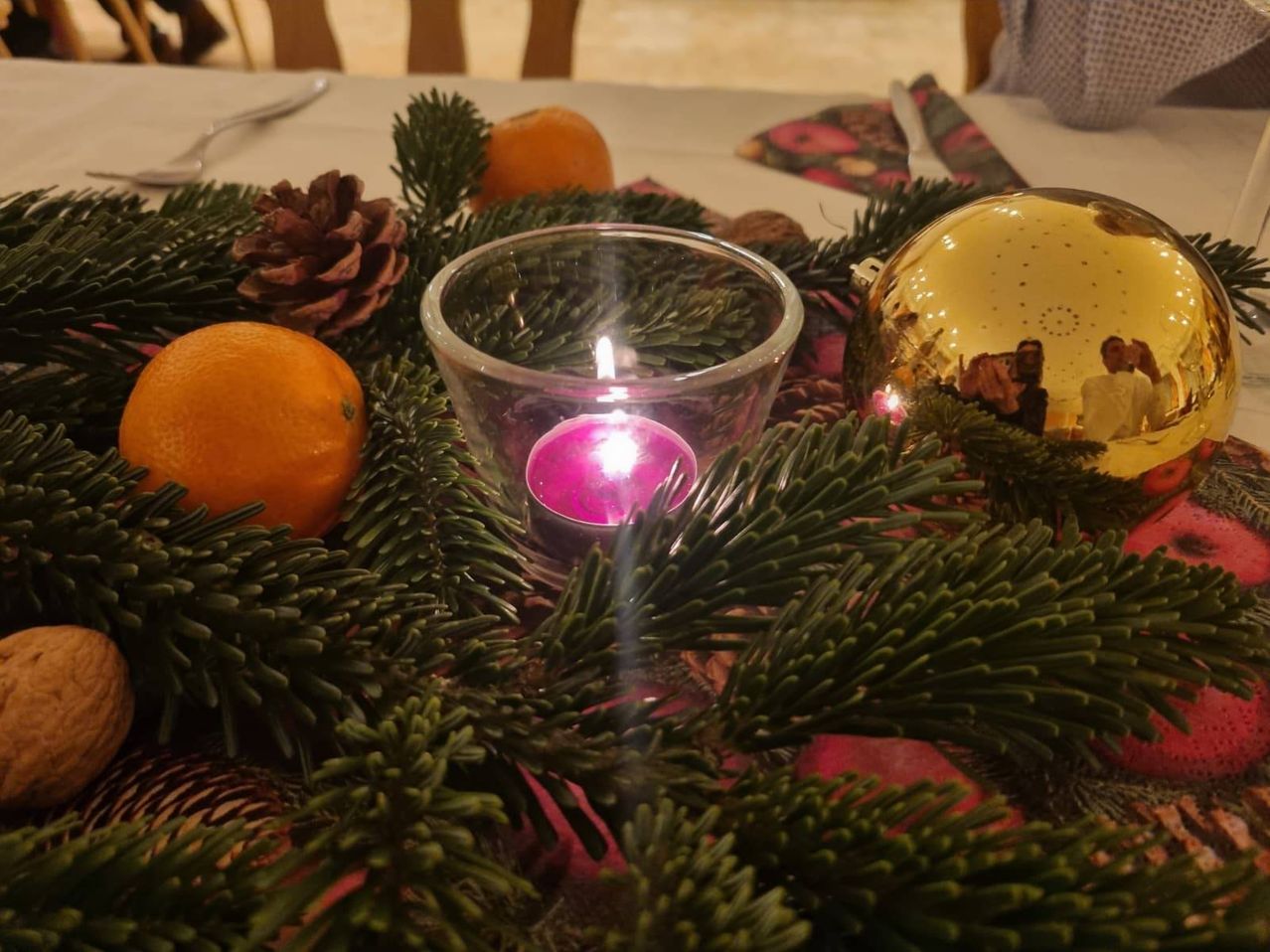 Ein Tisch ist mit Tannenzweigen, Orangen und einer brennenden Kerze geschmückt, wobei eine goldene Kugel Menschen im Hintergrund reflektiert.