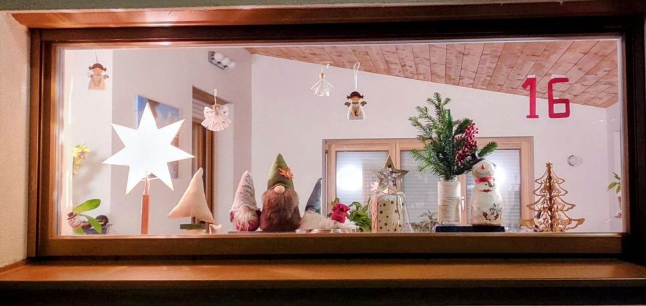 Ein Holzregal zeigt Dekorationsartikel, darunter Zwerge, Ornamente, einen Stern und Blumen in einem Vasen, mit einem Fenster, das eine Decke und Lichter reflektiert.