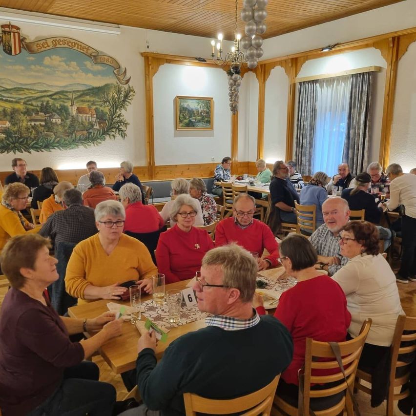 Eine Gruppe von Menschen sitzt in einem Esszimmer um Tische herum, einige tragen Brillen und Hüte. Ein Banner hängt an der Wand und eine Kronleuchter hängt von der Decke.
