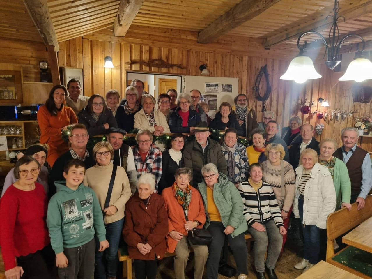 Eine Gruppe von Menschen versammelt sich drinnen, einige sitzen und andere stehen, mit festlichen Dekorationen und warmer Beleuchtung, lächeln für ein Gruppenfoto.