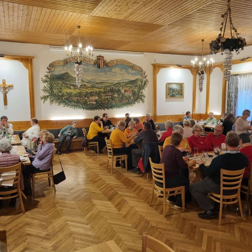 Eine Gruppe von Menschen sitzt an Tischen in einem Restaurant. Eine Wand ist mit einem Wandbild eines Dorfes geschmückt, und ein Kreuz ist darüber angebracht. Holzstühle und -tische sind überall angeordnet.