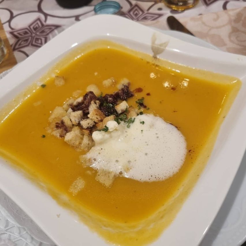 Ein quadratisches weißes Schüsselchen mit gelber Suppe, überzogen mit einem Klecks Sahne, Kräutern und gerösteten Brotkrumen.