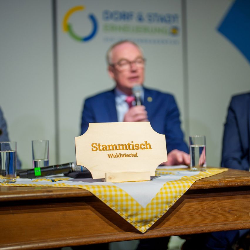 Ein Tisch mit einer gelb-weiß karierten Tischdecke, einem Mikrofon und zwei Gläsern Wasser. Ein Mann sitzt am Tisch, hält ein Mikrofon und spricht. Ein Schild auf dem Tisch steht Stammtisch Waldviertel.