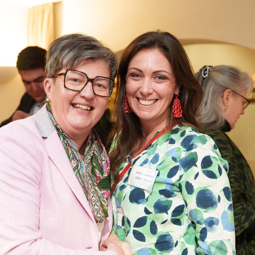 Zwei Frauen, eine mit Brille und Schal, die andere mit roten Ohrringen und Namensschild, lächeln und stehen bei einer Veranstaltung eng beieinander.